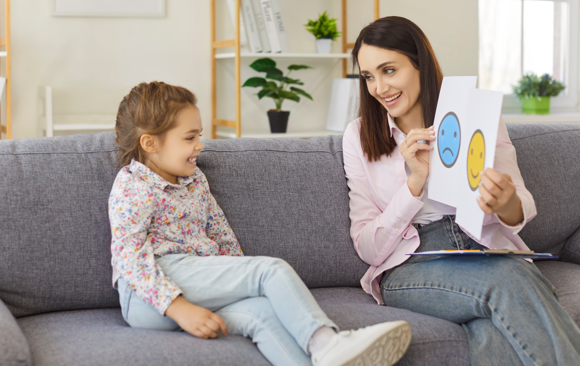 Psychologist and child test social skills, understanding language of emotions on meeting and therapy session in office. Young woman showing cards with sad and happy faces to kid. ADHD, autism concept